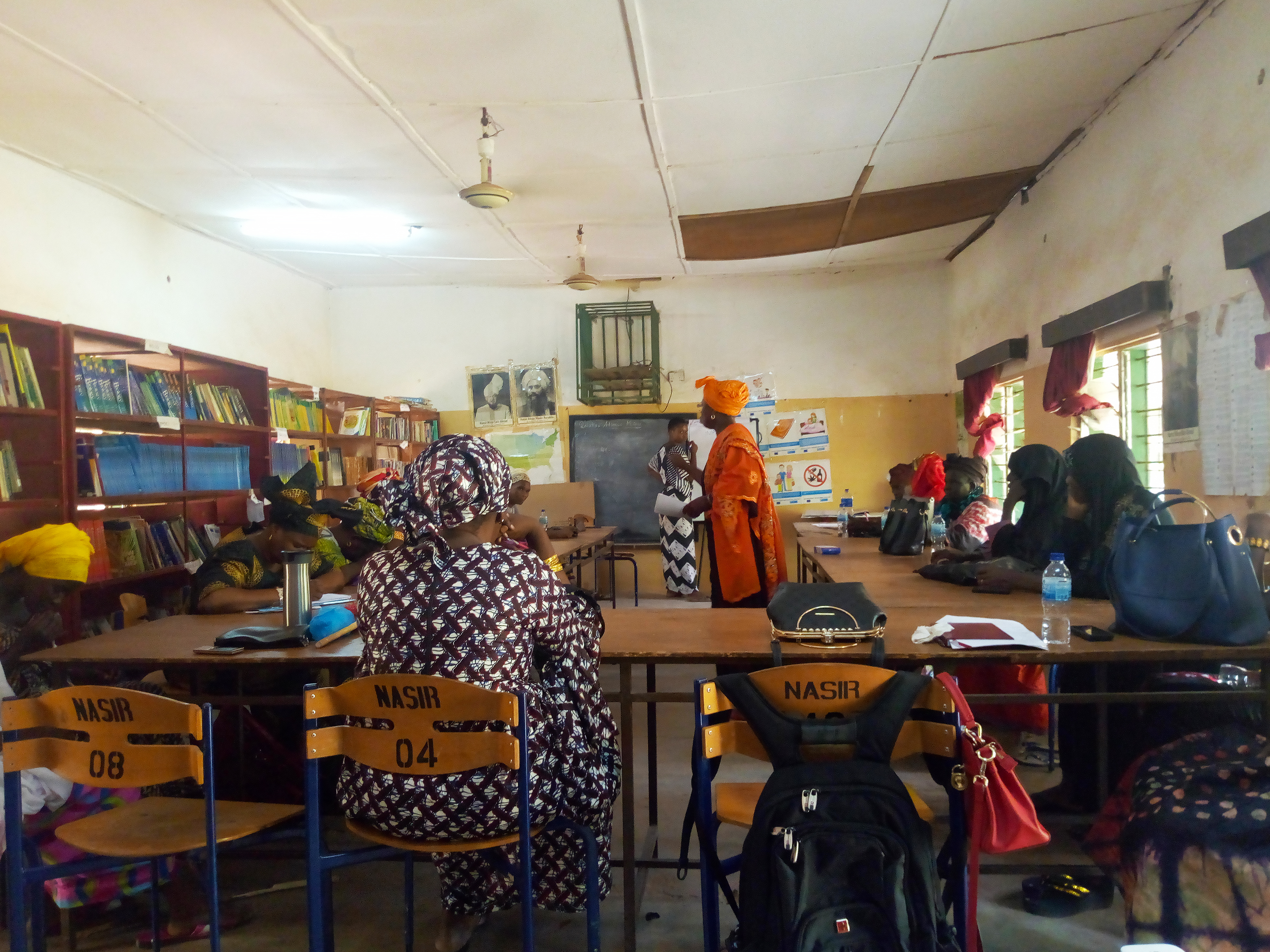 Women in The Gambia meeting in a classroom, sitting at school desks and writing on a black board.