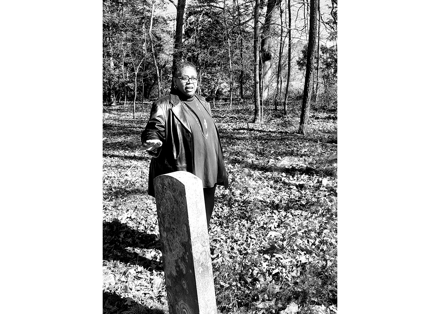 In black and white, a Black woman stands gesturing at a headstone in front of her, in a cemetery with grass and trees around her.