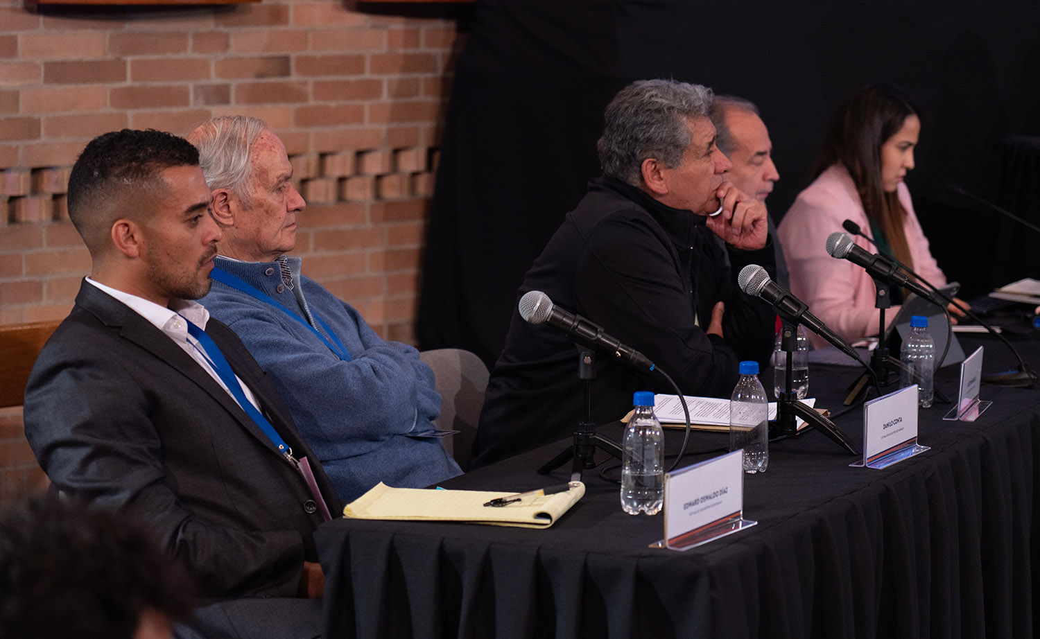 Five people sit at a table with microphones and papers in front of them facing the right hand side, listening.