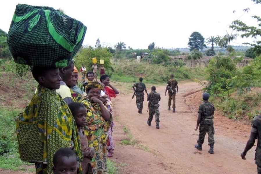 La mujer y los niños se paran en el lado izquierdo, viendo a los soldados armados pasar por un camino de tierra.