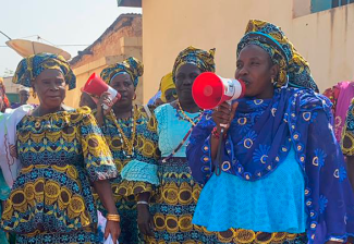 Several women in colorful traditional Gambian attire stand outside in the daylight, a couple of them holding megaphones