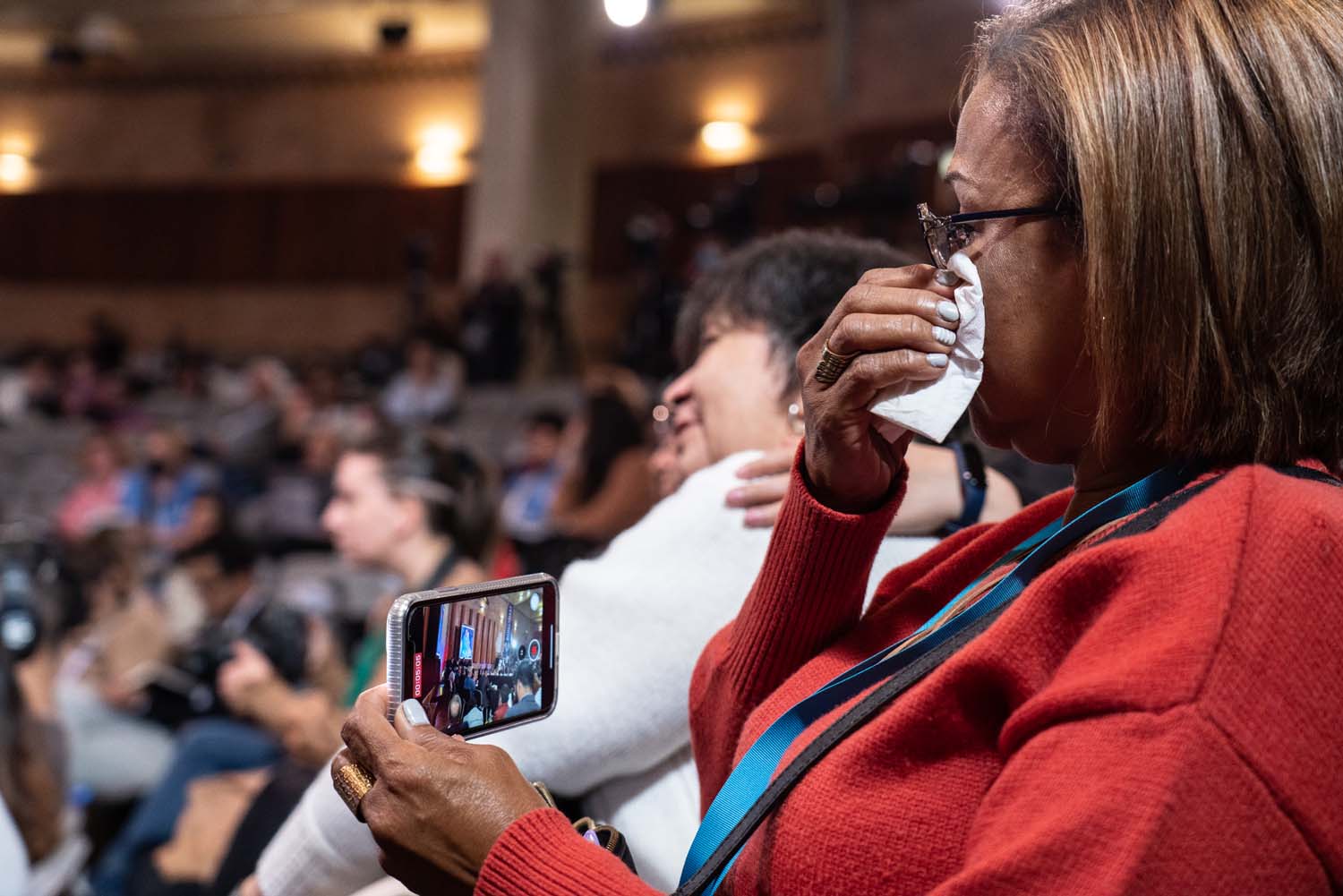 Una mujer se sienta en la audiencia, graba la audiencia en su teléfono celular y se limpia las lágrimas con un pañuelo.