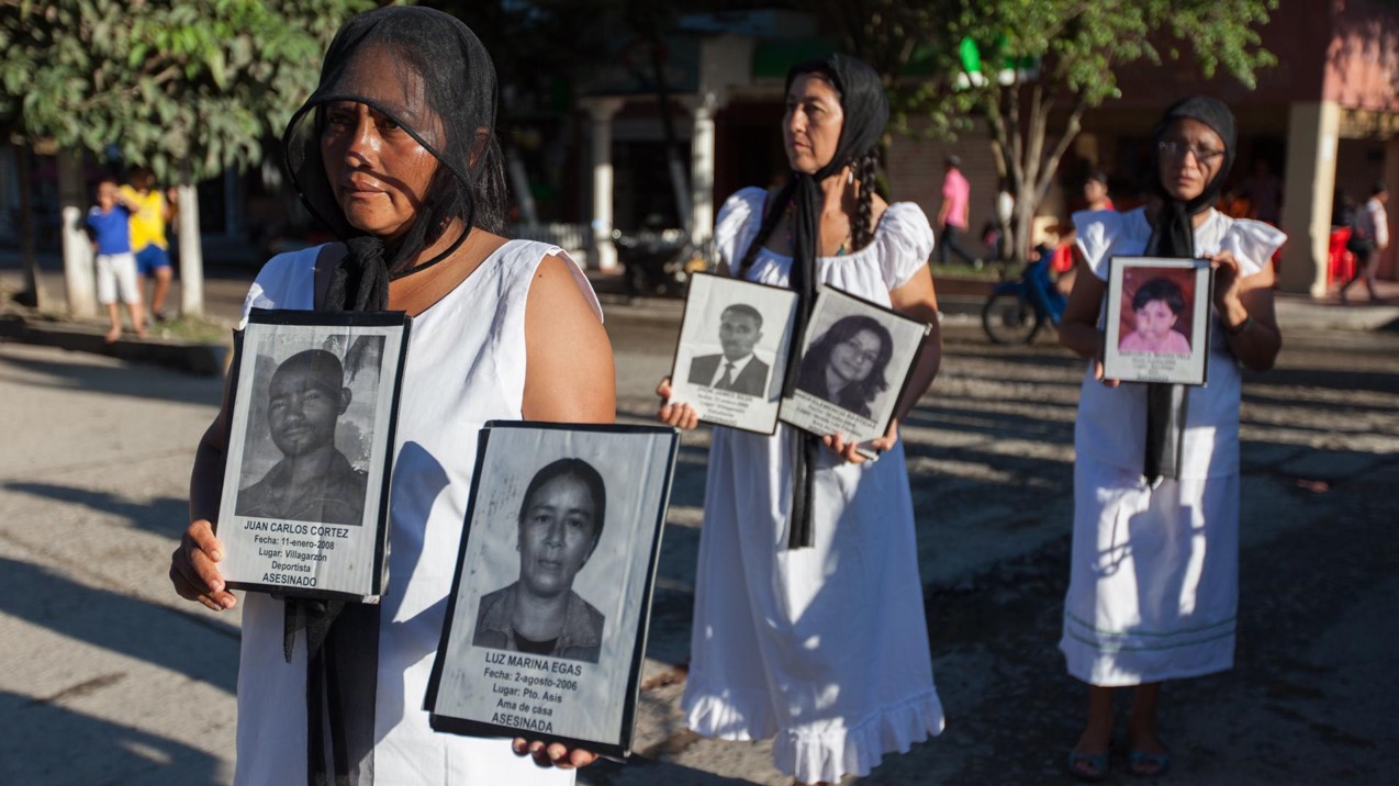 Three Latina women stand in a line, wearing white dresses, black headscarves, and all three are holding photographs of people