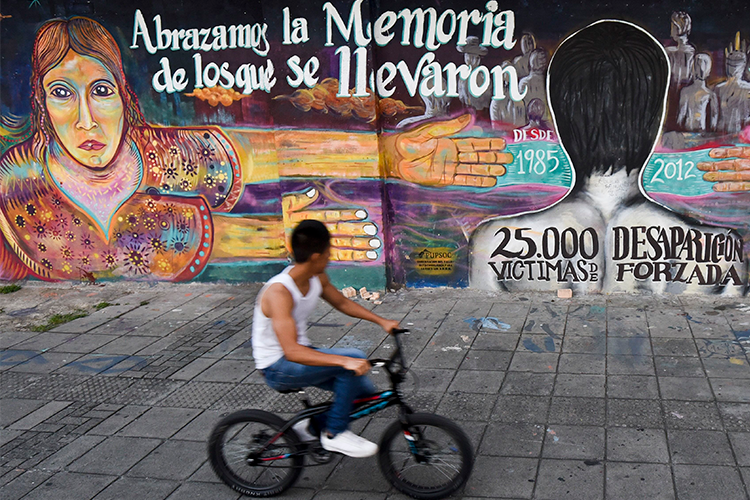 A man on a bicycle rides past a building whose exterior wall is covered in a colorful mural with writing in Spanish. 